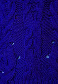 Gros plan sur un tissu tricoté bleu vif avec des motifs de torsades tressées et de petits trous ouverts laissant apparaître un fond bleu clair.