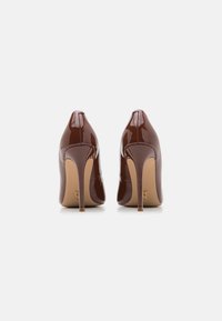 Un par de zapatos de tacón alto brillantes de color marrón oscuro vistos desde atrás sobre una superficie blanca, con tacones de aguja y puntas afiladas.