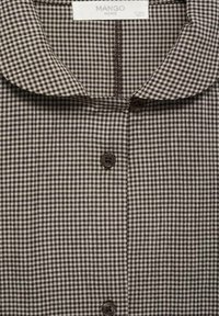 Camicia a quadri marrone e bianca con colletto ripiegato, chiusura con bottoni e tessuto testurizzato. Le etichette indicano il nome della marca e l'origine.