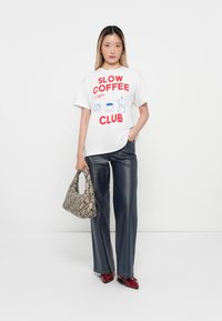 Balta kokvilnas T-kreklā ar sarkaniem un ziliem "SLOW COFFEE CLUB" grafikiem, apvienota ar tumši ziliem plaša piegriezuma biksēm un rakstainu bežīgu somu.
