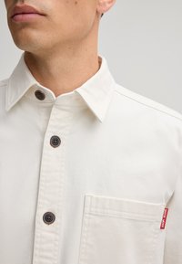 Beige overhemd met knopen en een kraag, met donkerbruine knopen, een borstzak en een klein rood merklabel op de zak.