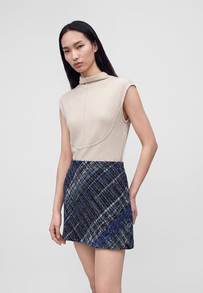 Femme aux cheveux noirs et lisses portant un haut beige sans manches et une mini-jupe à carreaux bleue, debout devant un fond clair uni.