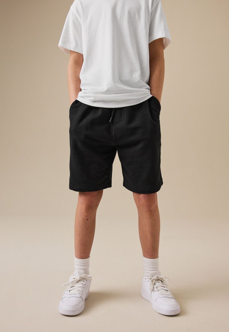 Jeune personne portant un short noir, un t-shirt blanc, des chaussettes blanches et des baskets blanches, debout, les mains dans les poches, sur fond beige.