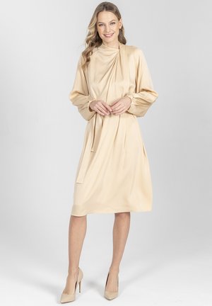 Kobieta w beżowej sukience z długim rękawem do kolan z drapowanym dekoltem, stojąca i uśmiechająca się, w pasujących szpilkach na jasnym tle.