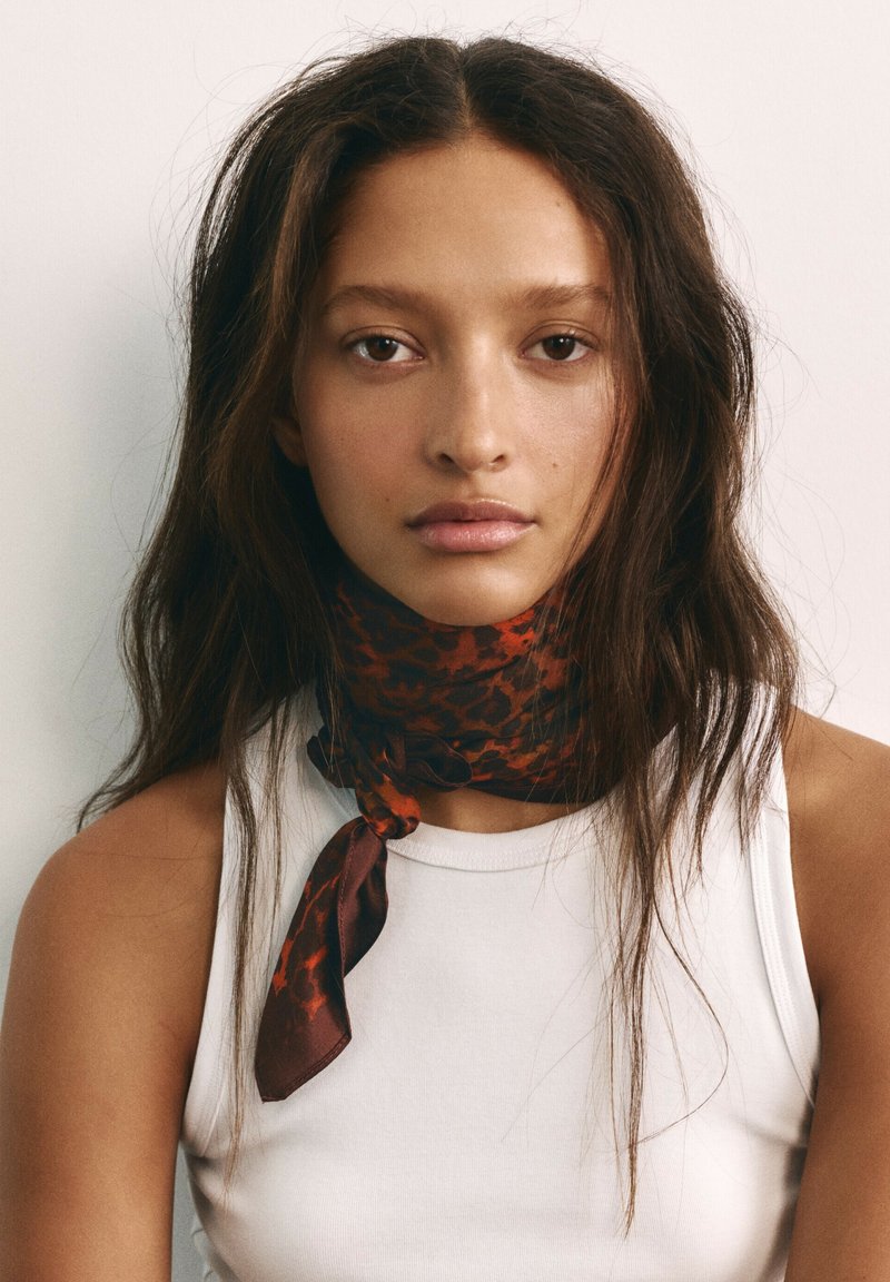 Jeune femme aux longs cheveux ondulés porte un top blanc sans manches et une écharpe marron et noire à motifs nouée autour du cou, regardant droit devant elle.