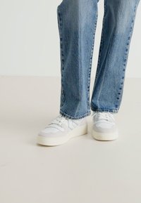 Hvite og lys grå sneakers har en teksturert overflate, med en polstret såle og flere snøringhull, sammen med blå denimjeans.