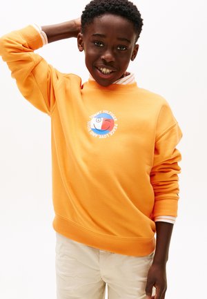 Χαμογελαστό αγόρι που φοράει πορτοκαλί φούτερ με λογότυπο ειρήνης και αγάπης, με το ένα χέρι πίσω από το κεφάλι, στέκεται μπροστά σε ένα απλό λευκό φόντο.