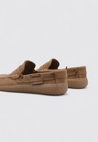 Zapatos de cuero marrón con una textura suave, diseño sin cordones, costuras decorativas y suela de goma; presentan un acento de cordón y detalle de logo.