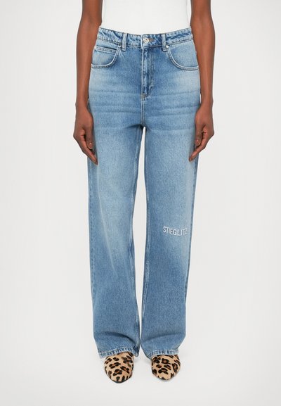 Jeans en denim bleu clair à jambes larges, dotés de cinq poches et d'un détail brodé "STIEGLITZ" sur la jambe gauche. Texture lisse, design décontracté.