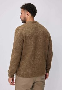 Homme aux cheveux courts et bouclés portant un pull brun texturé et un pantalon beige, vu de dos sur un fond clair uni.
