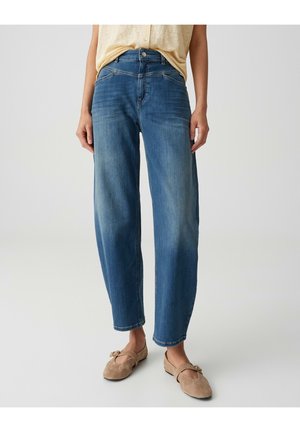 Frau trägt hochgeschnittene, locker sitzende blaue Jeans mit ausgewaschenen Details, kombiniert mit beigen flachen Wildlederschuhen mit Schnallenriemen.