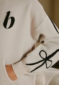 Lys beige sweater lavet af blødt stof, med et stort sort bogstav 'b' på brystet og et sort sløjfedesign på ærmet.