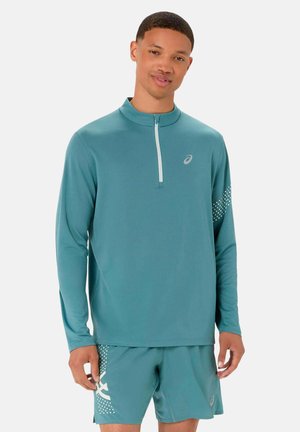 Giovane uomo che indossa una maglia sportiva a maniche lunghe color turchese abbinata con zip frontale e pantaloncini, in piedi davanti a uno sfondo neutro.