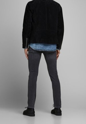 Person wearing a black jacket, blue denim shirt, dark gray jeans, and black shoes standing facing away against a plain background.