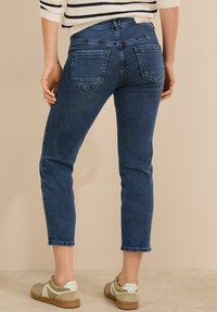 Jean en denim avec une taille mi-haute, des jambes fuselées, un lavage bleu foncé et trois poches arrière ; portés avec des baskets de couleur claire sur un fond neutre.