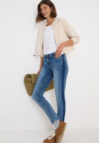 Lätt beige jacka över en vit t-shirt, kombinerad med blå jeans som har en mörkblå sidstripe. Bruna sneakers fullbordar outfiten.