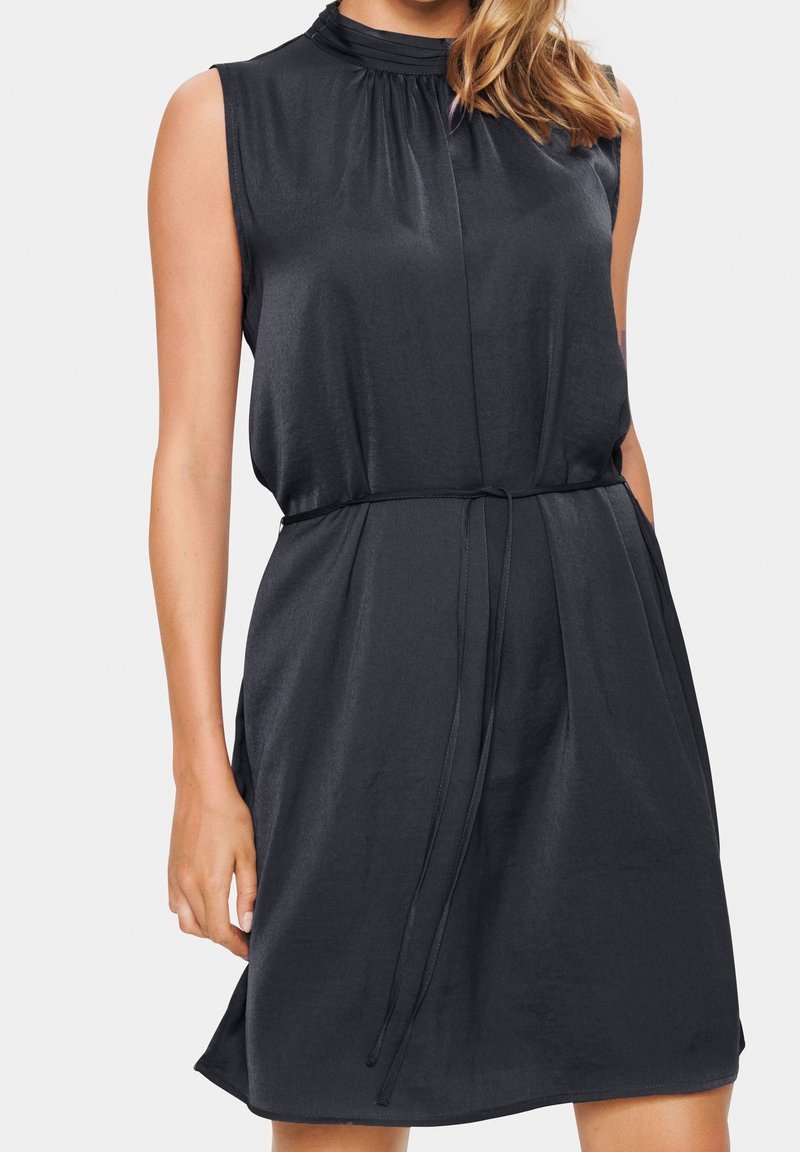 Robe noire en satin sans manches avec un col haut froncé et une fine ceinture à la taille, portée par une personne aux cheveux blonds lâchés.