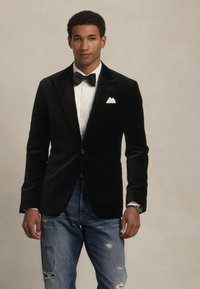 Črna žametna blazer jakna s pikčastimi reverji, oblečena čez belo srajco in metuljčkom, v kombinaciji z utrujenimi modrimi trapericami. Vidna naglavna roba.