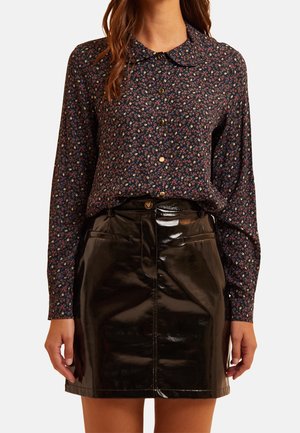 Vrouw draagt een donkerblouse met lange mouwen en bloemenprint, ingestopt in een glanzende zwarte mini-rok van lakleer met voorzakken en knoopsluiting.