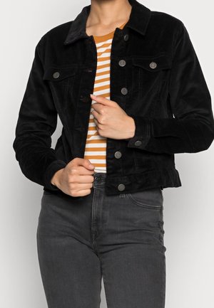 Personne portant une veste noire à boutons par-dessus une chemise rayée blanche et orange et un jean gris foncé, ajustant la veste à la taille.