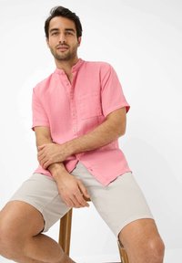 Camicia di lino rosa a maniche corte con colletto alla cinese, dotata di taschino sul petto e dettagli con bottoni, indossata con pantaloni corti beige chiaro.