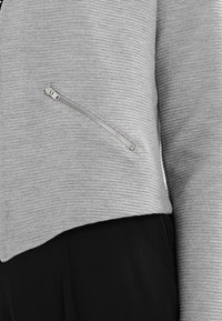 Szara skórzana kurtka z krótkim rękawem o żebrowanej teksturze, z detalem zamka błyskawicznego w kieszeni oraz eleganckim krojem. Materiał wydaje się miękki i lekki.