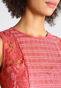 Röd spetsblus med en strukturerad, vävd mönstring. Halsringningen är rund med en kontrasterande slät kant, som visar upp intrikata spetshöjd på sidan.