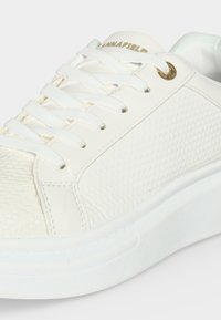 Λευκό χαμηλό αθλητικό παπούτσι με πλεκτή υφή στην πλαϊνή επιφάνεια, λευκά κορδόνια και χρυσή λεπτομέρεια στη τρύπα κοντά στον αστράγαλο.