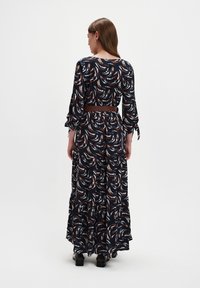 Kobieta w długiej, ciemnej sukience w wzory z rękawami do łokci i brązowym paskiem, stojąca tyłem na jednolitym białym tle.