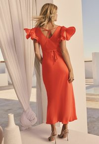 Robe midi rouge avec un décolleté à nouer, des manches à volants et un tissu lisse. Portée avec des chaussures à lanières de ton neutre sur un fond clair.
