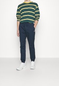 Grön- och gulrandig långärmad skjorta; marinblå byxor med snörning; vita sneakers; avslappnad passform, med logotypdetaljer på skjortan och byxorna.