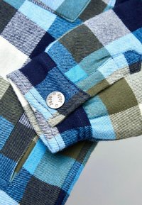Camicia di flanella con un motivo a quadri blu, verde e bianco. Dettaglio a bottoni sul polsino con finitura argentata. Texture morbida.