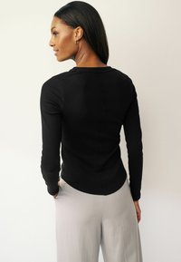 Black long-sleeve top with a fitted design, ribbed texture, subtle seam detail along the back, paired with light grey wide-leg pants.