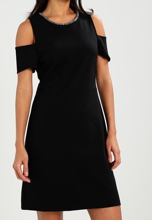 Black knee-length dress with cold shoulder cutouts and silver beaded neckline detail, worn by a standing woman.