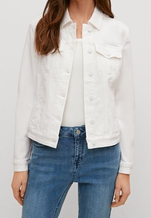 Femme portant une veste en jean blanche boutonnée sur un haut blanc et un jean bleu, montrée des épaules jusqu'aux hanches.