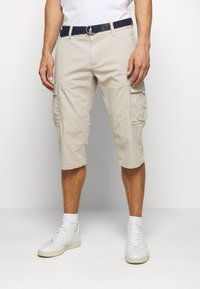 Shorts de cargo beige con dos bolsillos laterales, material de algodón, hasta la rodilla, combinados con un cinturón azul marino y zapatillas blancas. Textura suave.