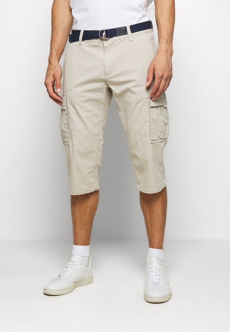 Shorts de cargo beige con dos bolsillos laterales, material de algodón, hasta la rodilla, combinados con un cinturón azul marino y zapatillas blancas. Textura suave.