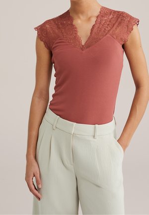 Frau trägt ein ärmelloses, rostfarbenes Oberteil mit Spitzenärmeln und hellbeige, hoch taillierte, plissierte Hose, Hände entspannt seitlich.