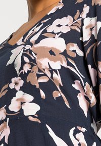 Top floreale realizzato in tessuto morbido, con base blu navy e fiori rosa e beige. Scollo a V e maniche fluide con una texture liscia.