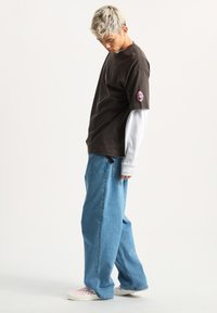 Camiseta de color marrón oscuro con un gráfico rosa en el lado izquierdo del pecho, superpuesta a una camiseta de manga larga gris claro, combinada con un vaquero azul holgado.