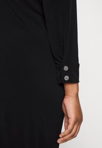 Manica di un vestito nero con tessuto liscio, design annodato alla spalla e bottoni decorativi vicino al polsino con iniziali incise.