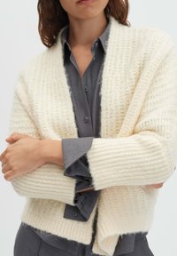 Donna che indossa un cardigan a maglia grossa crema sopra una camicia grigio scuro con bottoni, braccia incrociate all'altezza della vita, inquadratura tagliata dal naso ai fianchi.