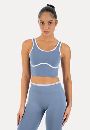 Kobieta stojąca, ubrana w niebieski, dopasowany zestaw prążkowanego sportowego crop topu i legginsów z wysokim stanem z białymi wykończeniami.