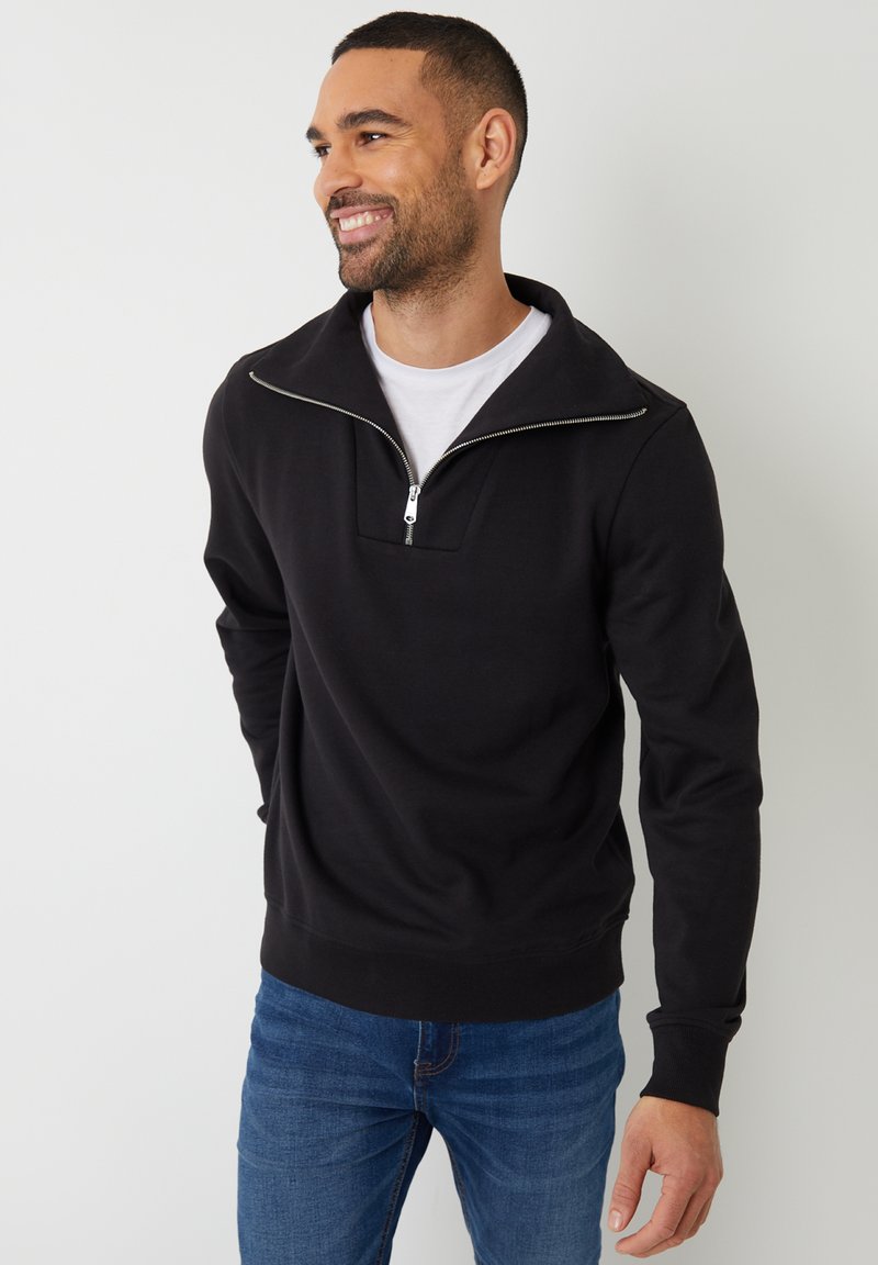 Zwarte ritssluiting sweater met hoge kraag, gemaakt van zacht materiaal. Heeft een vierkante halslijn en geribde manchetten, gedragen over een witte t-shirt.