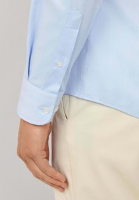 Muška ruka pored džepa krem hlača, nosi svijetloplavu košulju s dugim rukavima i dugmadi na manžetama.