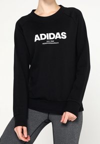 Sweat-shirt noir avec logo blanc "ADIDAS" et texte "EST. 1949 HERZOGENAURACH". Dispose de poignets et d'un col côtelés avec une coupe décontractée.