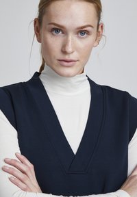 Top senza maniche di colore navy con scollo a V profondo, indossato sopra un dolcevita bianco. Tessuto liscio, design minimalista e dettagli di cucitura sottili.