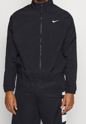 Mann trägt schwarze leichte Jacke mit Reißverschluss und weißem Nike-Logo auf der Brust sowie schwarze Hose mit weißem Streifen an der Seite.
