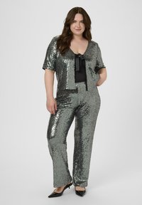 Ensemble deux pièces pailleté en argent et noir. Veste à manches courtes avec un détail en nœud, pantalon ajusté avec une texture brillante, et chaussures noires à bouts pointus.