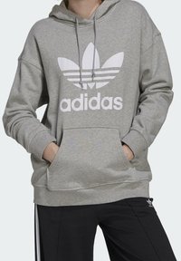 Grå hoodie med en stor vit Adidas-logga och tre ränder på bröstet. Har en dragsko i huvan och en känguruficka. Svarta byxor med sidskaror.